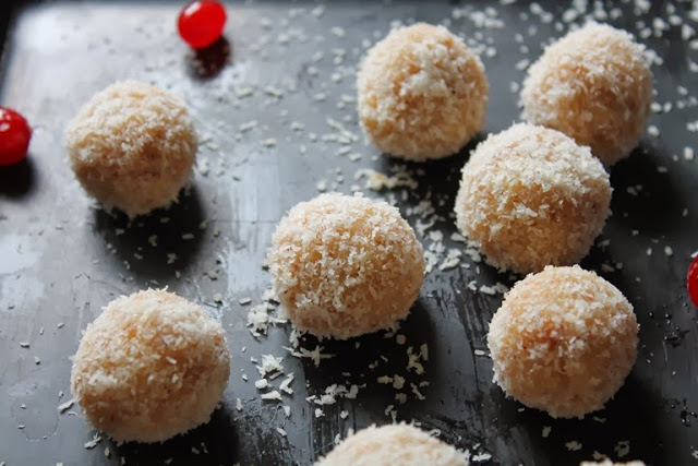 coconut ladoo displayed on a black serving dish scattered with desiccated coconut and red cherries