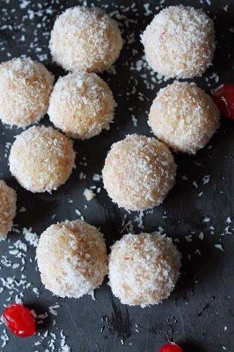 coconut ladoo displayed on a black serving dish scattered with desiccated coconut and red cherries