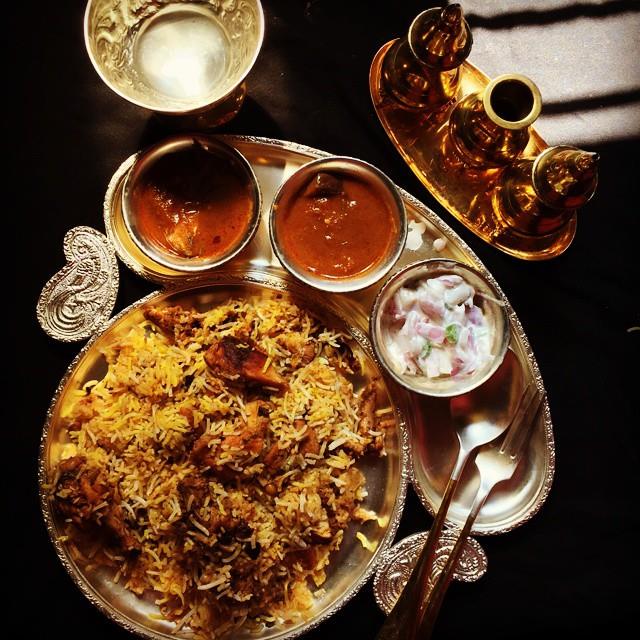 chicken dum biryani served on a brass plate with onion raita, biryani brinjal gravy & salan