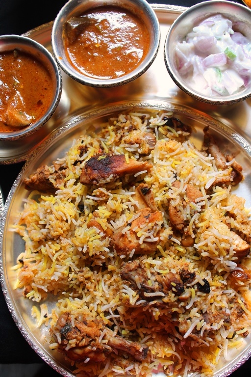chicken dum biryani served on a brass plate with onion raita, biryani brinjal gravy & salan