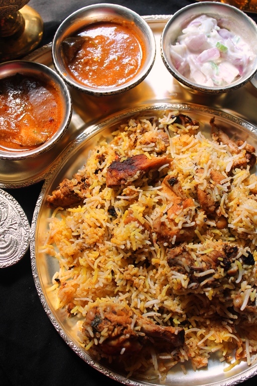 chicken dum biryani served on a brass plate with onion raita, biryani brinjal gravy & salan