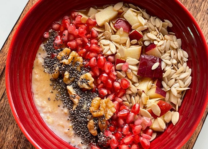 oats porridge served in a red bowl with chia seeds, walnuts, pomegranates, melon seeds, chopped apples and pumpkin seeds.