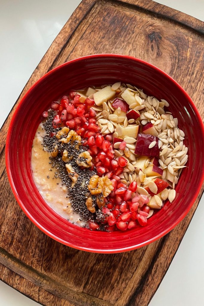 oats porridge served in a red bowl with chia seeds, walnuts, pomegranates, melon seeds, chopped apples and pumpkin seeds.