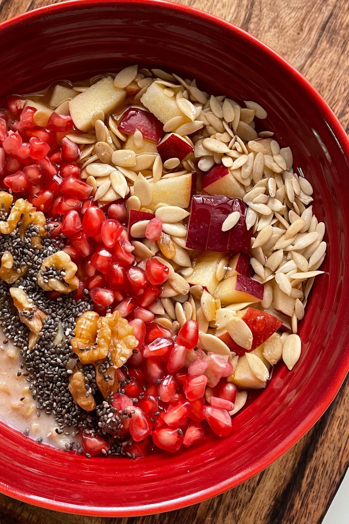 oats porridge served in a red bowl with chia seeds, walnuts, pomegranates, melon seeds, chopped apples and pumpkin seeds.